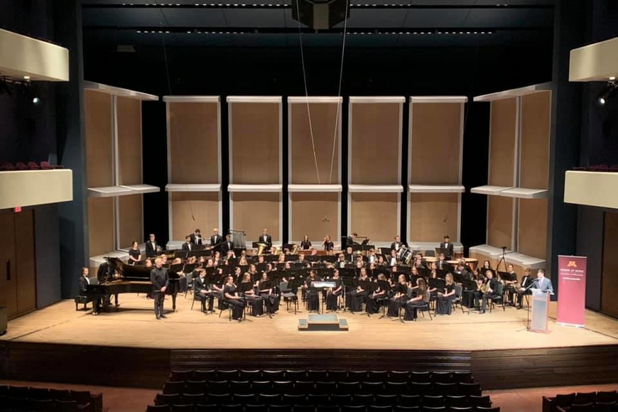 Concert band members seated and dressed in concert black on the Ted Mann Concert Hall stage.