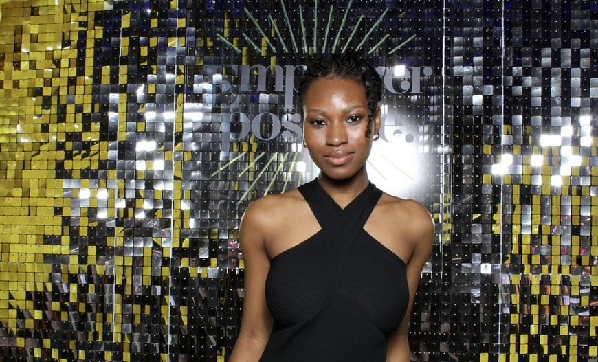 A black woman with black braids in a bun wearing a black dress, smiling in front of a black background with gold sequences
