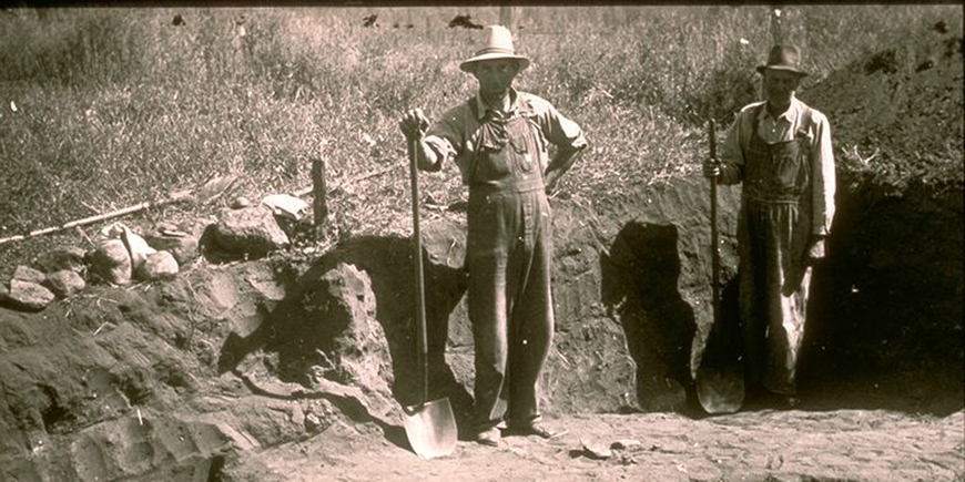 Black and white photo of two men wearing hats and holding a shovels