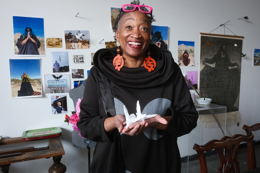 A smiling woman holds a small sculpture in her hands in front of photos tacked to a white wall behind her