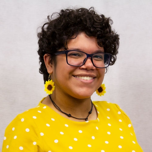 Photo of Maya, a biracial person with short curly hair, wearing a yellow dress.
