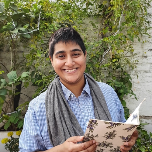 This is a photo of me, an Indian person with short hair, with a thin book in my hand sitting infront of a wall of green vines. I am wearing a grey scarf and a light blue shirt. 