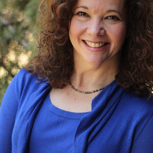 head shot of a white woman with dark curly hair, smiling, standing outside, wearing blue