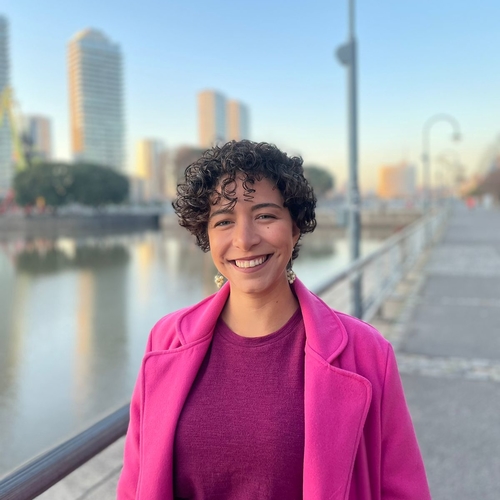 Larissa has short curly hair, olive/tan skin tone. She is wearing a purple blouse and a pink coat. She is smiling. The background is a river in Buenos Aires, there are some buildings and the sky is blue.