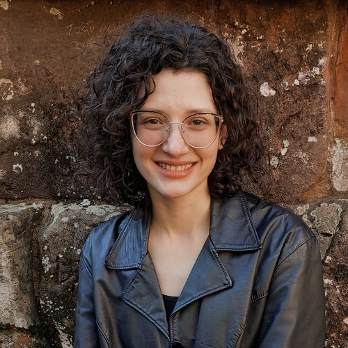A woman with light skin and medium-length curly brown hair, wearing glasses and a black jacket, smiles in the photograph.