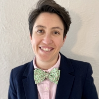 Michaeline's headshot. She is smiling, has short brown hair, and is wearing a navy blazer with a pink shirt and green bowtie.