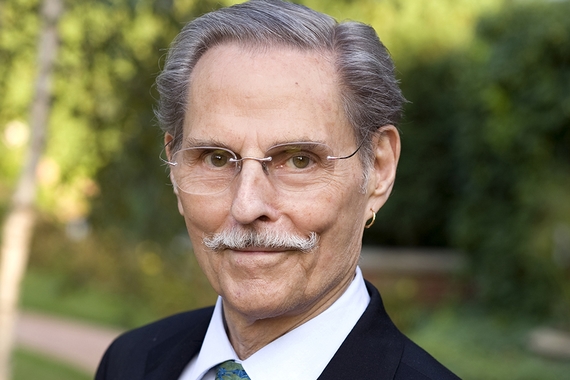 Head and shoulders of person with grey short hair and mustache and light skin, wearing wire-rimmed glasses and dark suit with white collar shirt