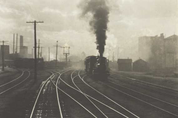 A black and white image of a train billowing smoke, there are many train tracks in the foreground