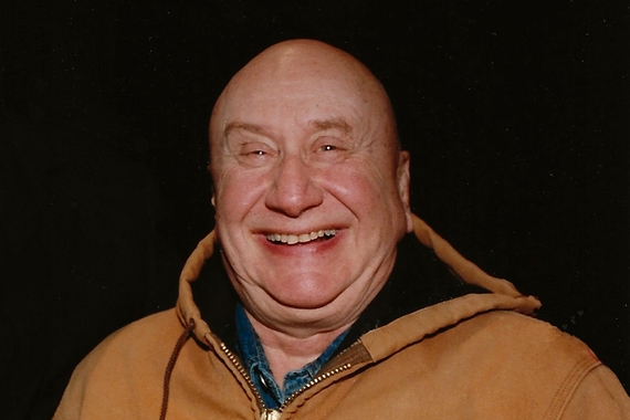 Head and shoulders of person with bald head and light skin, smiling and wearing a brown hooded jacket