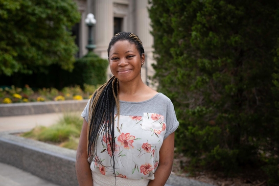 Smiling black girl standing outside in front of a pillared building and green trees. She wears a floral shirt with grey shoulder sleeves, white torso top sprinkled with pink florals, and white skirt. Has black and gold braids placed in a side sweep. 