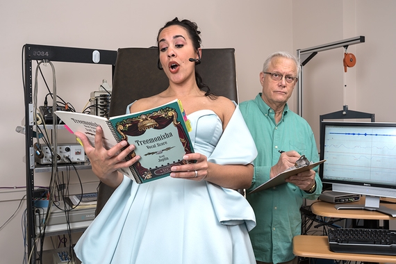 A woman is singing opera as a Dr. watches taking notes in a research lab.