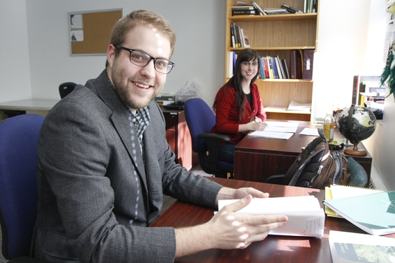Comm-Studies grad students sitting at separate desks smiling at camera