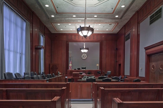 An empty courtroom that includes black chairs for the jury and lawyers, wood accents, white curtains over big windows, and the judge's seat