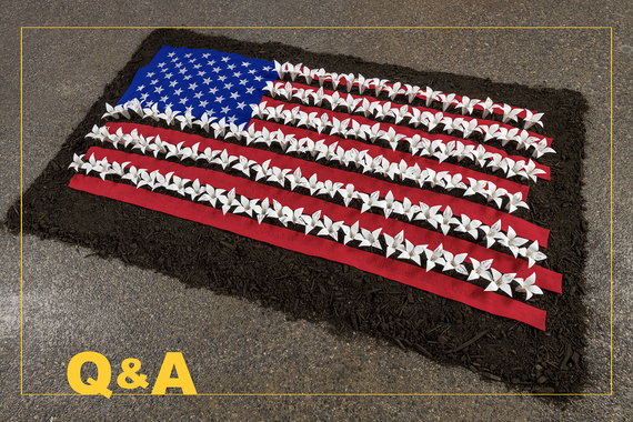 Art installation of American flag made of origami flowers on a pile of dirt on concrete floor.