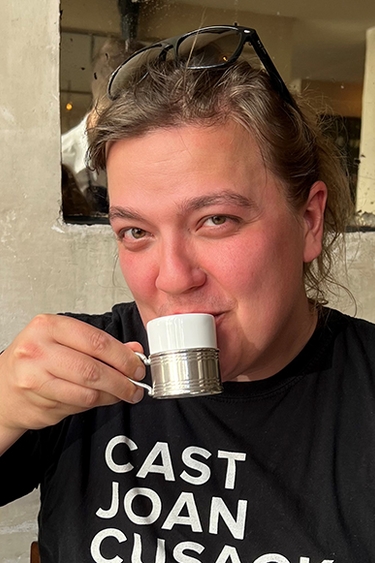 Head and shoulders of person sipping coffee; short brown hair, light skin, wearing black t-shirt with white text: Cast Joan Cusak