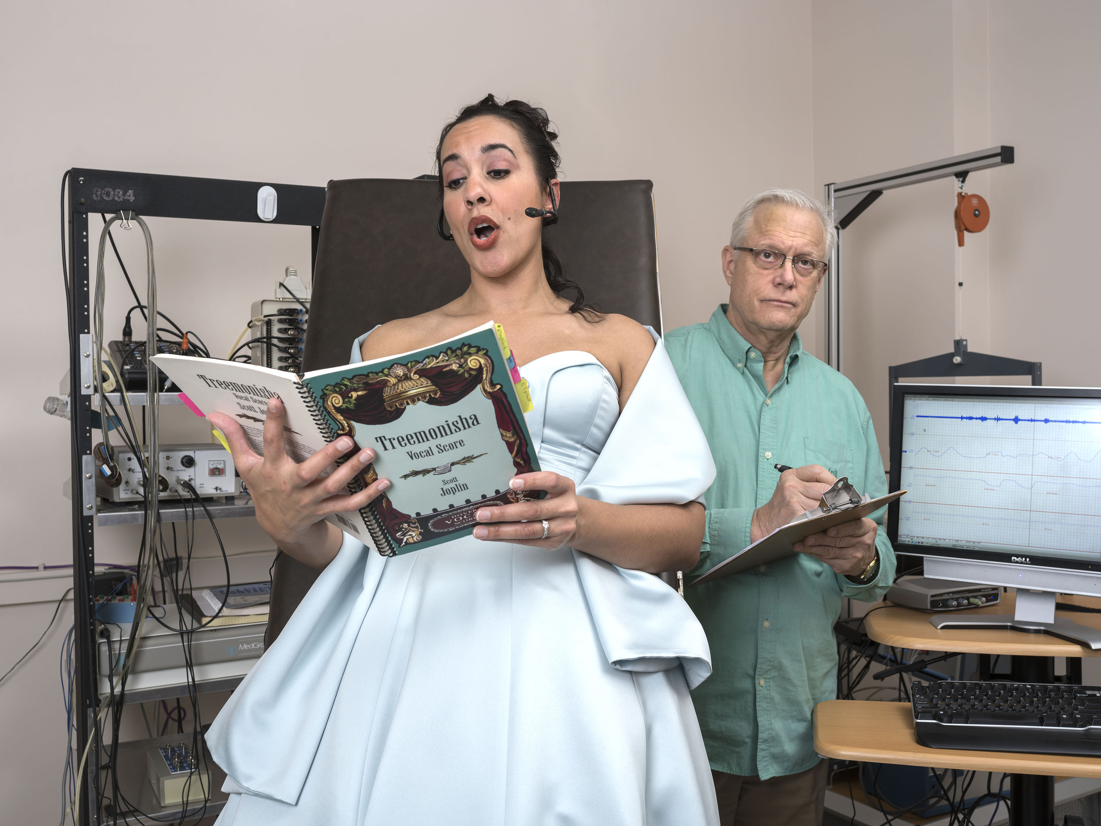 A woman is singing opera as a Dr. watches taking notes in a research lab.