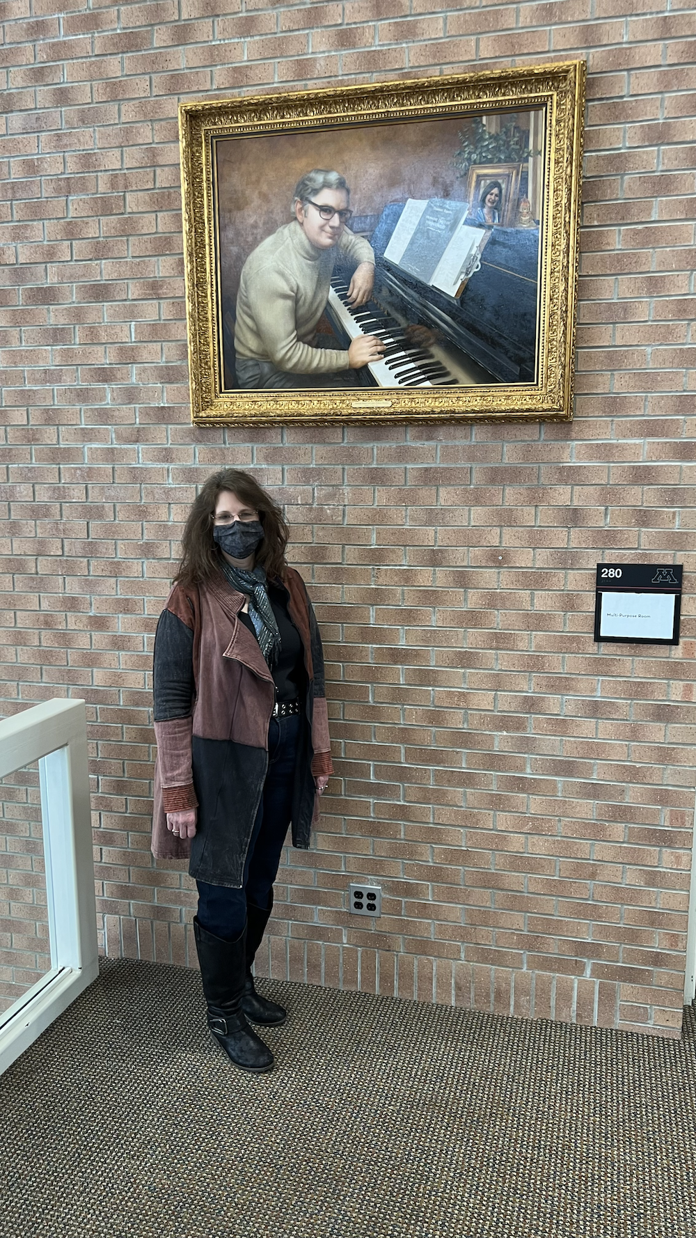 Artist Suzann Beck, wearing a black and brown patched coat and black pants, poses for a picture below Dominick Argento's portrait on the second floor of Ferguson Hall's lobby.