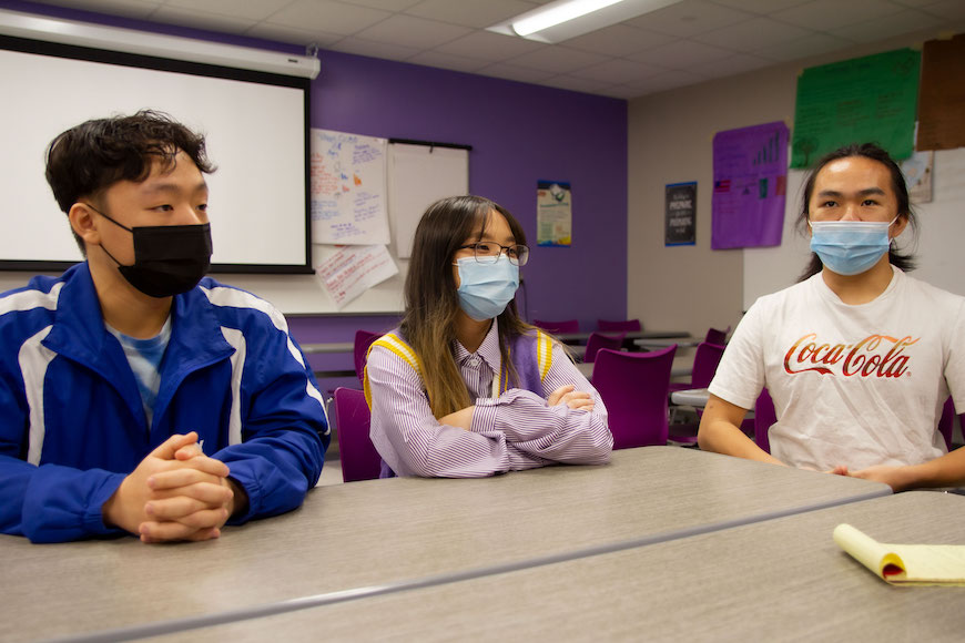 Three young people sitting at a table together in a high school classroom. They have dark hair, light brown skin and are wearing masks.