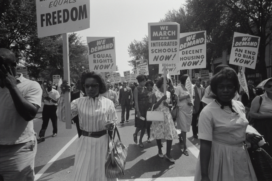 A procession of marchers carrying signs demanding freedom, equal rights, integrated schools, and decent housing.