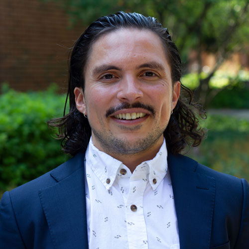 Dr. Juan Del Toro, a person with shoulder-length dark brown hair, a brown mustache, wearing a blue blazer and white button shirt