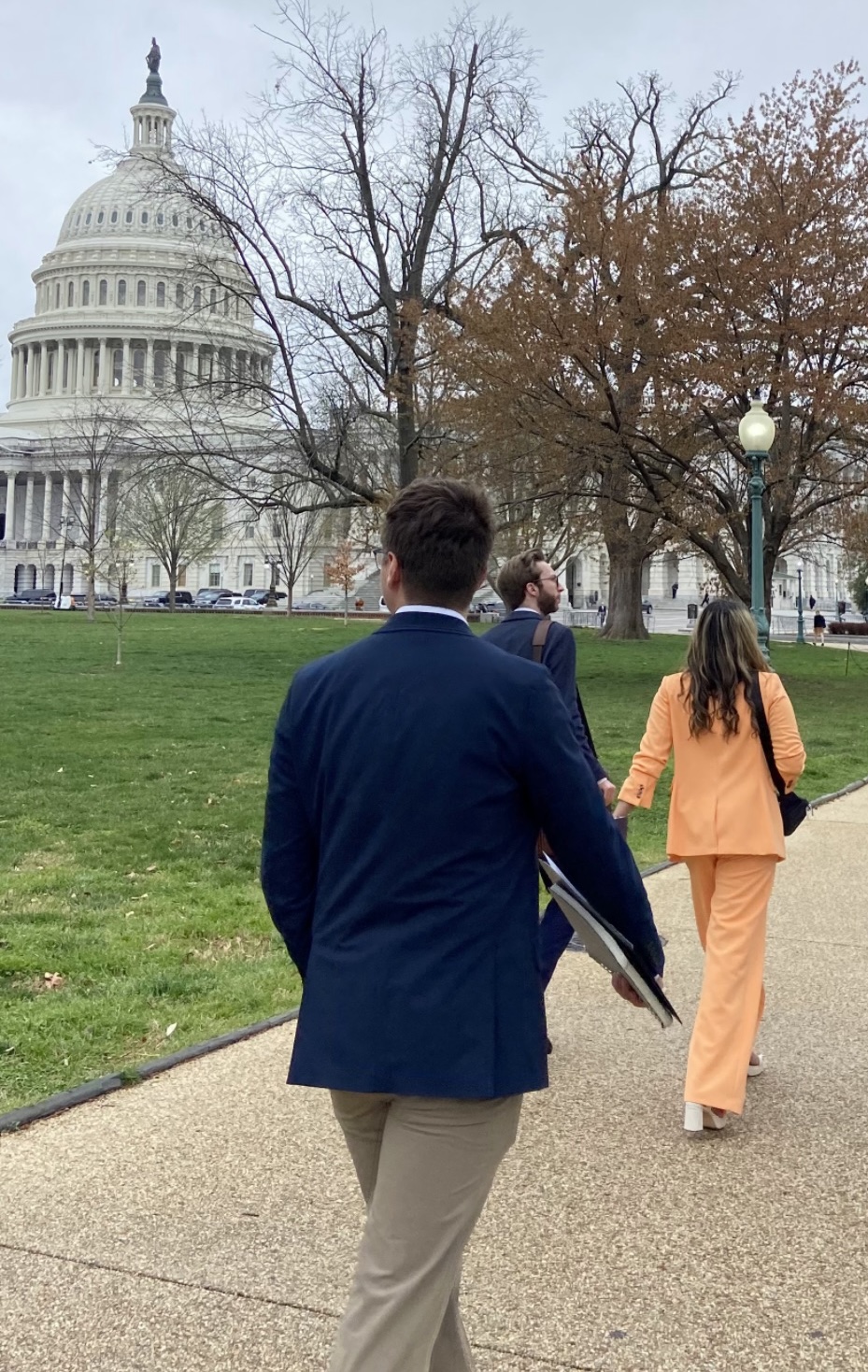 Hassan wears an peach colored suit and walks in front of the Nation's Capitol