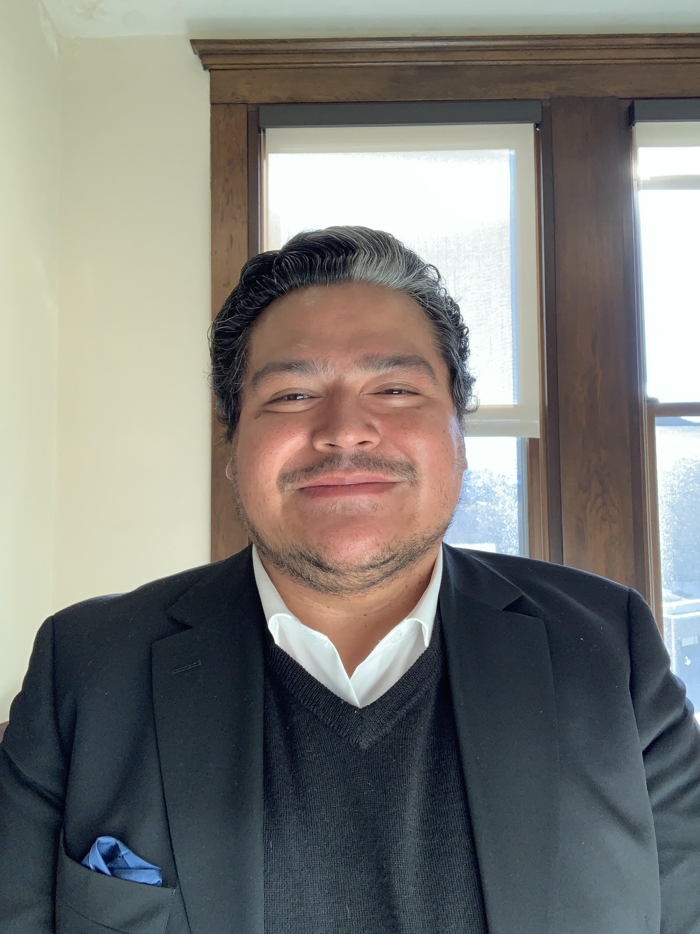 Osiris Aníbal Gómez, a person with short black and grey hair, light brown skin, smiling, wearing a jacket, sweater, and buttoned shirt, standing in front of a window