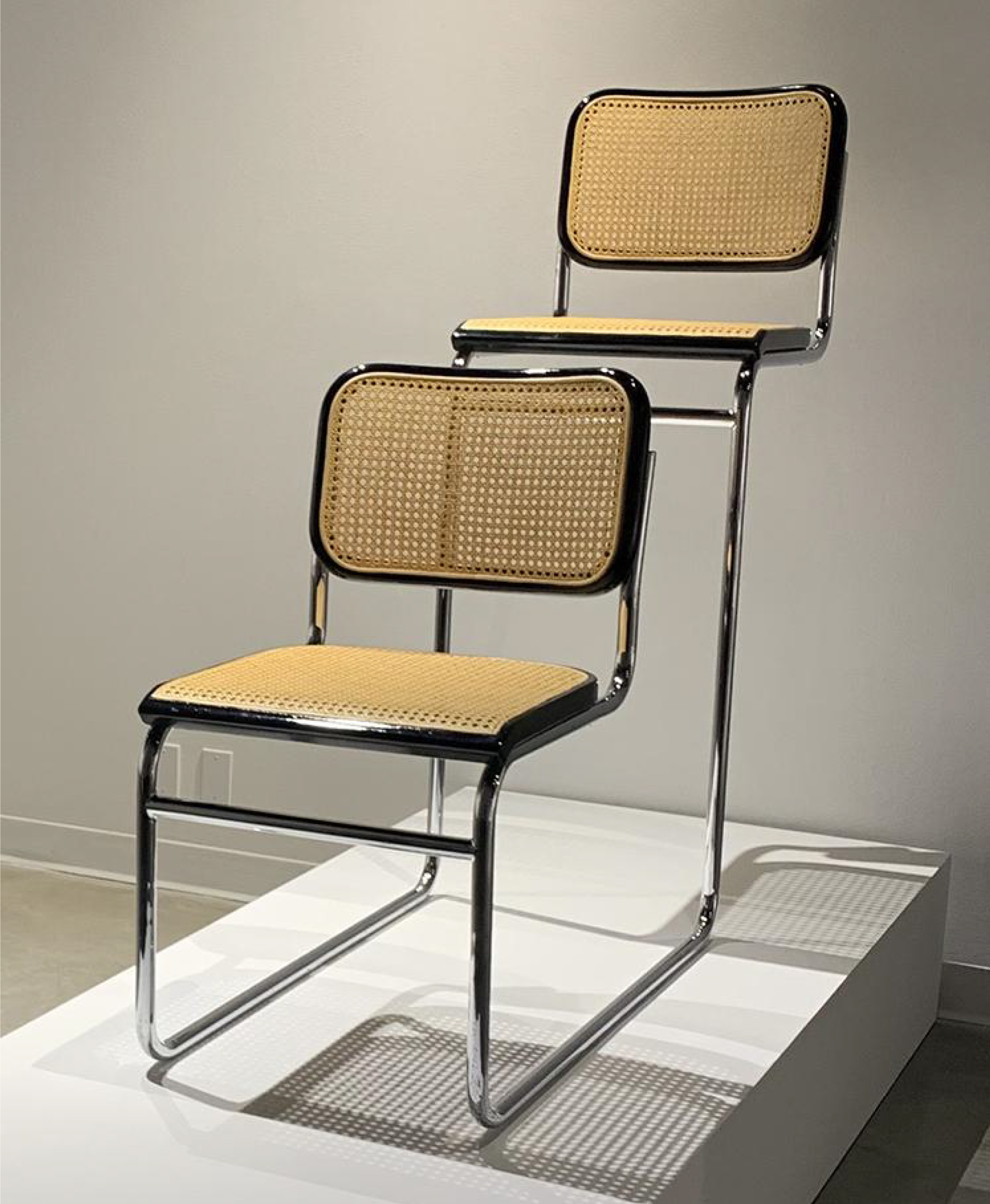 Two cane chairs connect by their chrome legs in a single file line with the chair in the front being smaller and the chair in the back being taller.