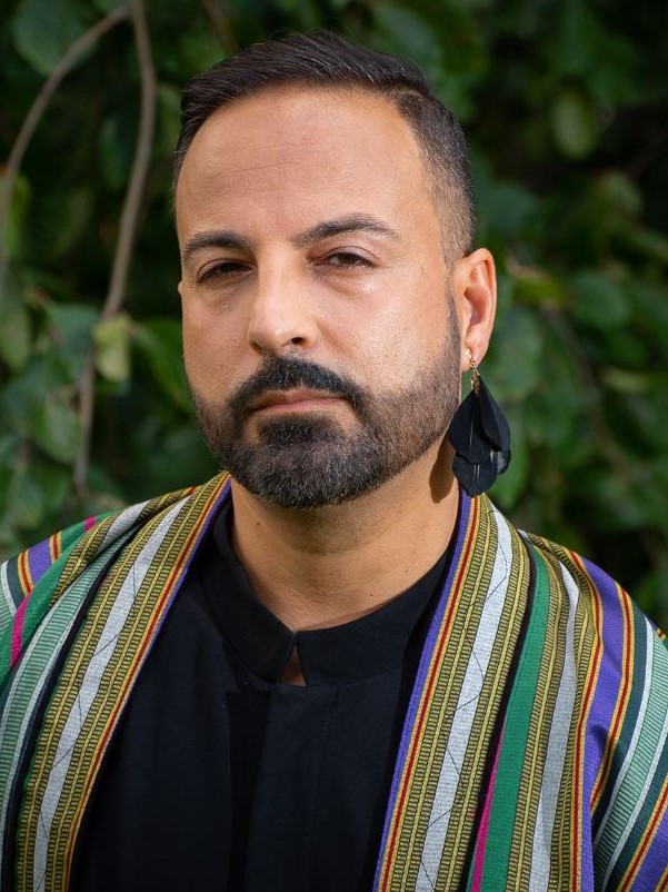 Headshot of Qais, wearing black shirt, multi-colored striped jacket, and black feather earring.