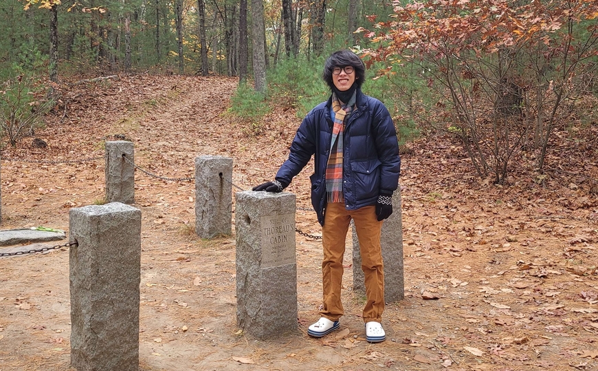 person standing by short stone pillars in autumn forest, person's hand is placed a pillar, person is tall, has straight black hair, tan skin, wears a thick blue coat, black gloves, auburn pants, white crocks, and striped scarf
