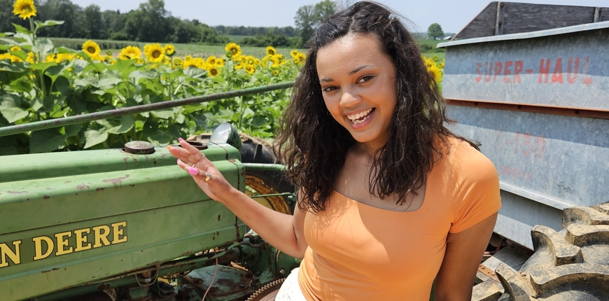 Head and torso of person with dark wavy hair to shoulders and light brown skin, smiling and wearing orange top and gestering at farm equipment and field of sunflowers