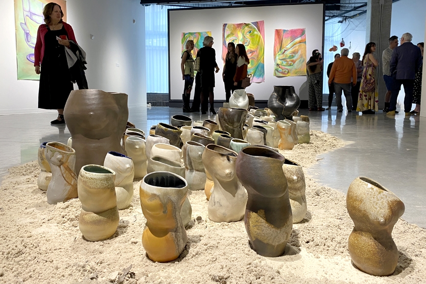 A collection of ceramic pots on a pile of sand on the floor of an art gallery with large paintings on the back walls and visitors milling about