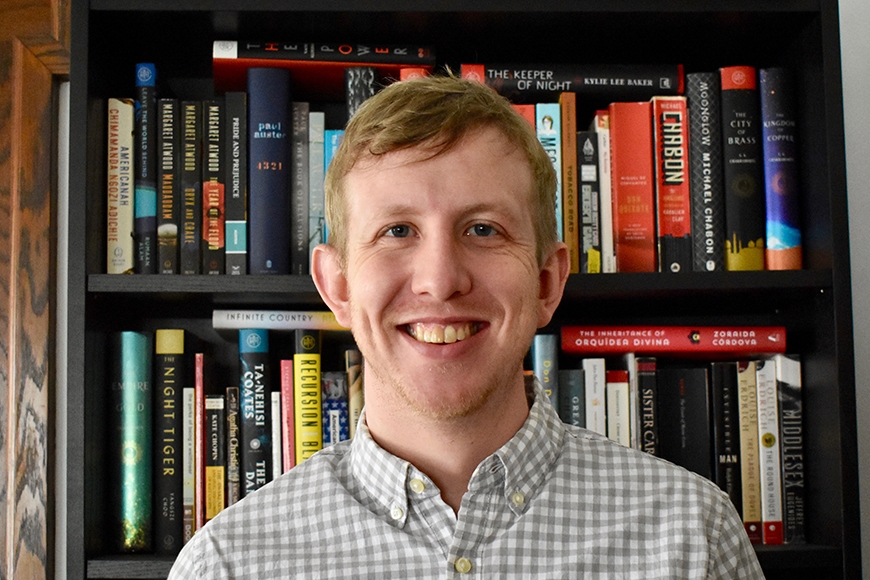 A smiling young man with short blond hair stands in front of a colorful bookshelf