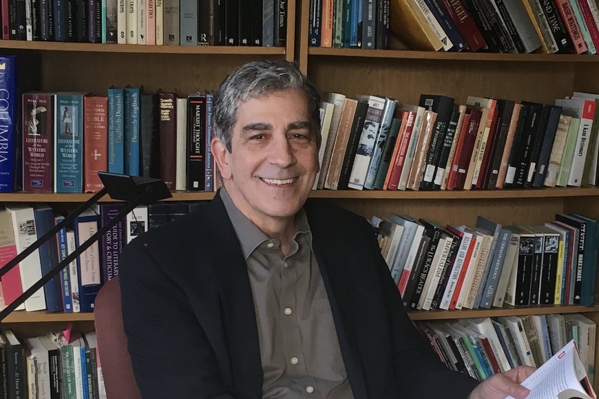 Head and torso photo of person with grey short hair and light skin, wearing dark blazer and grey button up shirt, holding book in left hand, sitting in front of bookshelves