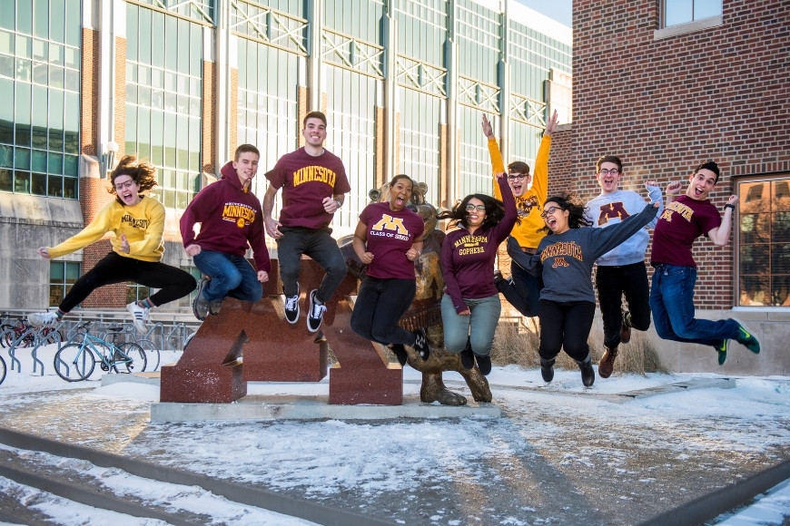 Several students wearing University of Minnesota gear cheering while jumping over a snowy platform.