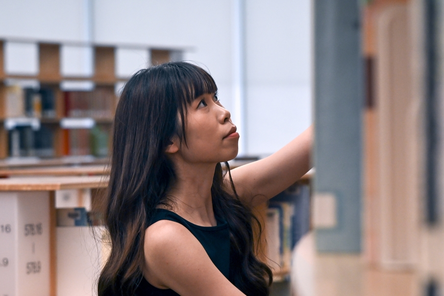 Person with long brown hair and bangs at eyebrow level looking and reaching for a book on a shelf above them. They are wearing a black tank top surrounded by other bookshelves.