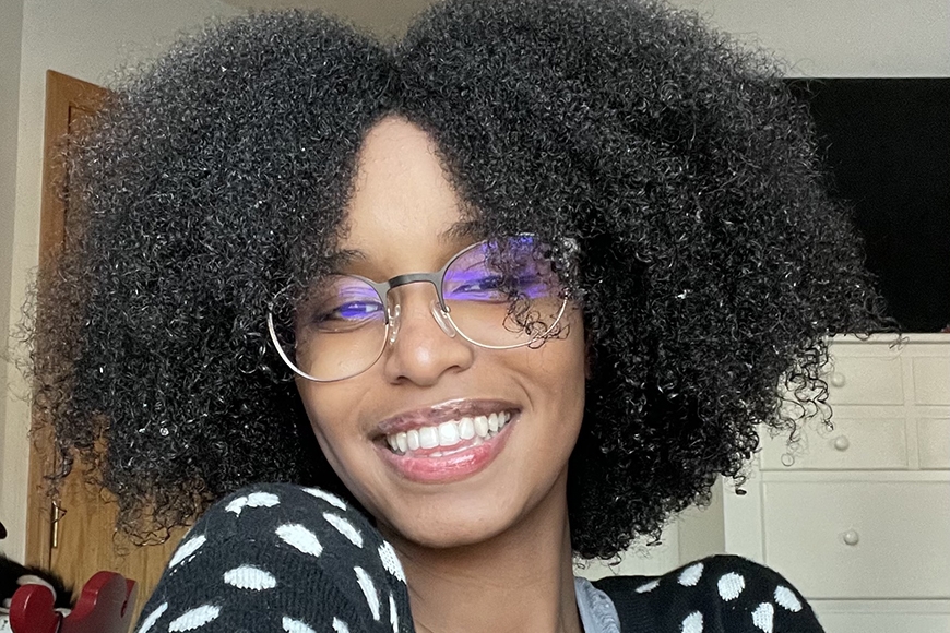 Head and shoulders image of person with black curly hair, light-rimmed glasses, light brown skin, smiling, wearing white polka dot black shirt, in front of white and black interior and brown door