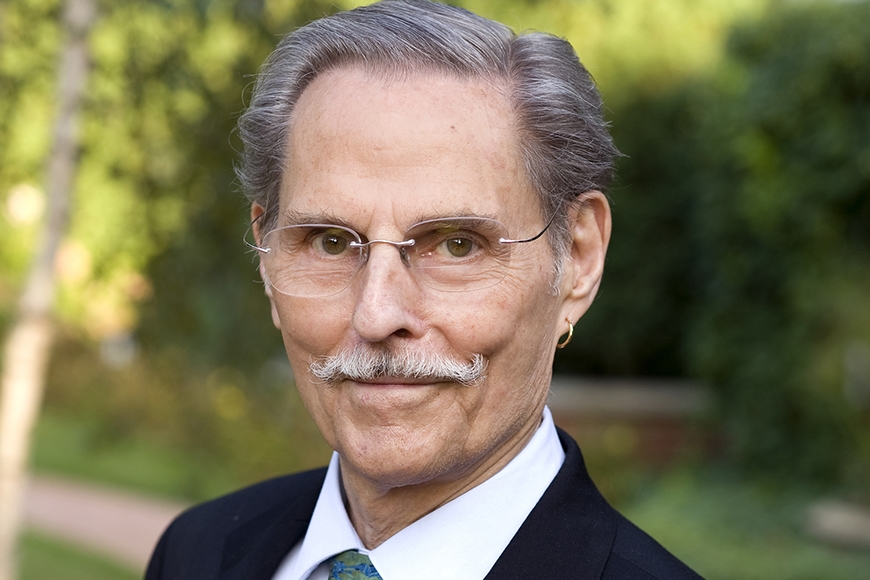 Head and shoulders of person with grey short hair and mustache and light skin, wearing wire-rimmed glasses and dark suit with white collar shirt