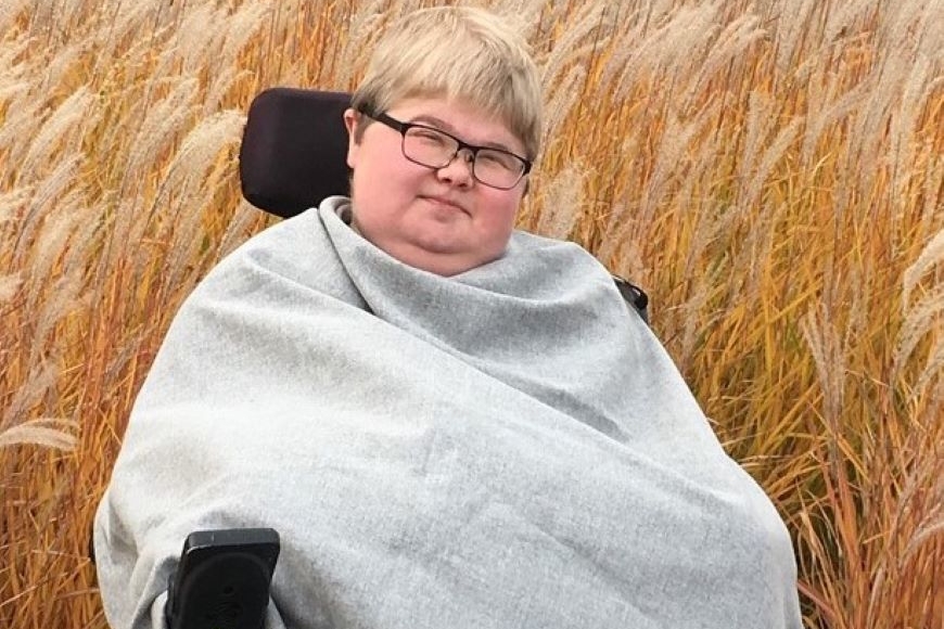 Cole Anderson, a person with short blond hair wearing glasses, seated in a wheelchair. In the background are tall grasses.