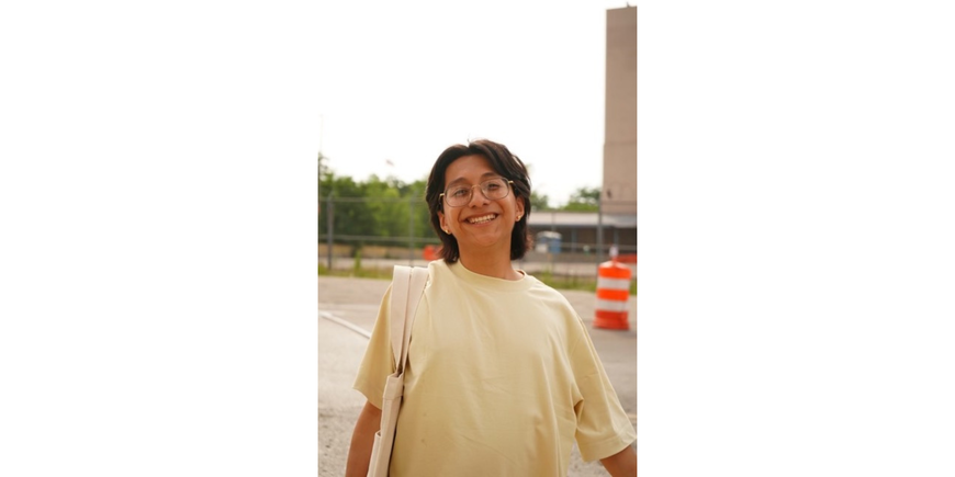 Portrait of a person with dark hair to shoulders, wearingthinly framed gold glasses and a light yellow t-shirt, smiling.