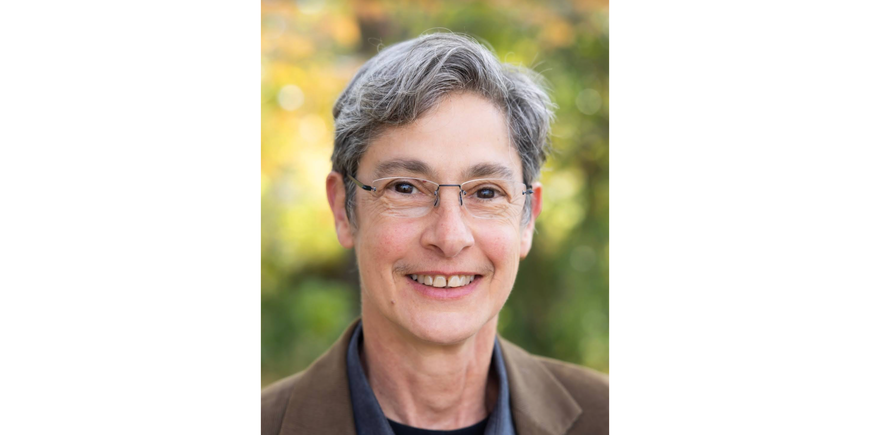 Portrait of a person with short grey hair, wearing thinly framed glasses, smiling. The person is standing in front of a green background while wearing a brown blazer and blue collared shirt. 