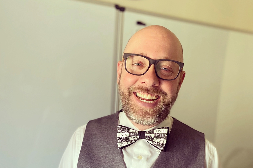 Head and shoulders photo of person with bald head, glasses, light skin, salt-and-pepper beard, smiling, wearing grey vest over white button shirt with grey and white bowtie in front of white background