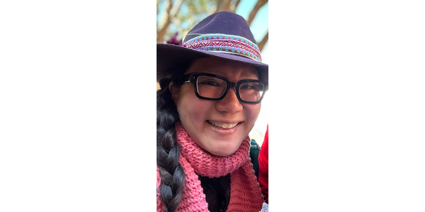 Portrait of a woman wearing black framed glasses and a grey hat with a short brim, smiling. The person is wearing a pink scarf wrapped around their neck and their hair is dark brown in a braid on their right side. 