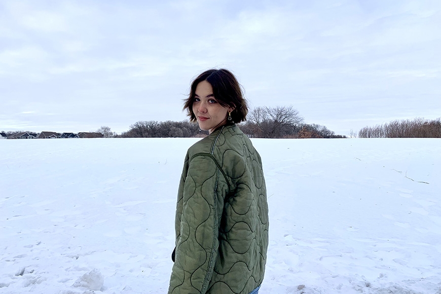 Head and torso of person standing in snow field with distant trees; hair to chin, earing green coat