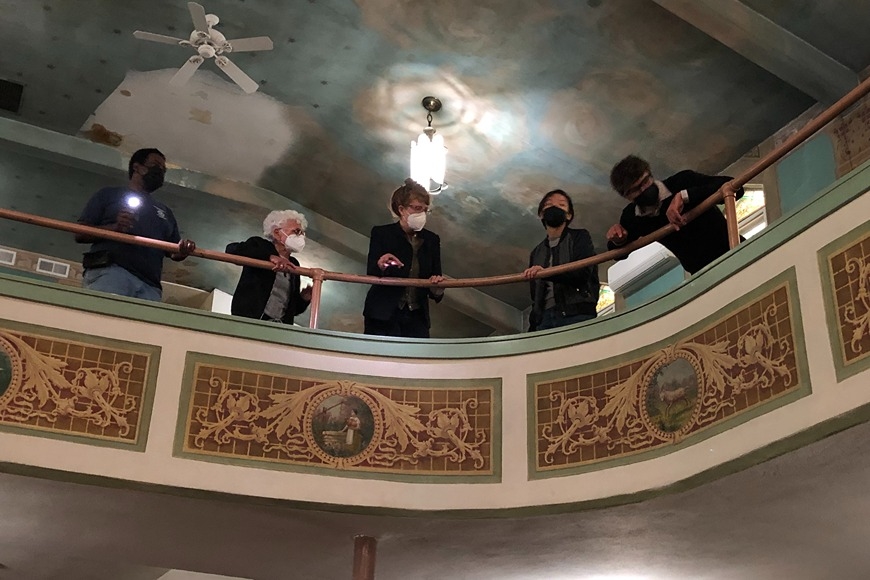 [From left to right] FCOGIC Director John Lewis, Dr. Marilyn Chiat, Jessie Merriam, Jade Ryerson, and Dr. Greg Donofrio standing atop a balcony in the FCOGIC building. The cielings are painted like the sky, and the face balcony painted with gold zodiac medallions