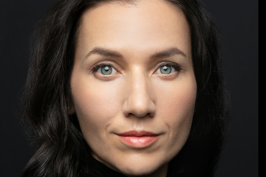 Head shot of person with dark hair past chin, light skin, wearing pink lipstick, in front of dark background