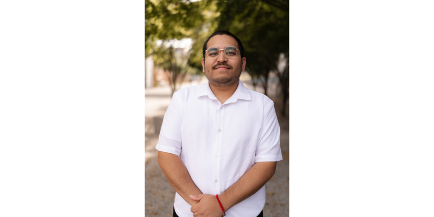 Person with short brown hair and a brown mustache standing with hands joined together smiling slightly. They are wearing a short sleeve white collared polo shirt and they are standing in front of a paved path with trees in the background.