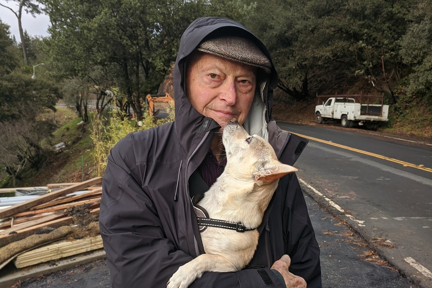 Head and torso of person with light skin wearing black hooded coat, tweed cap, and holding small white dog; in background trees and asphalt road