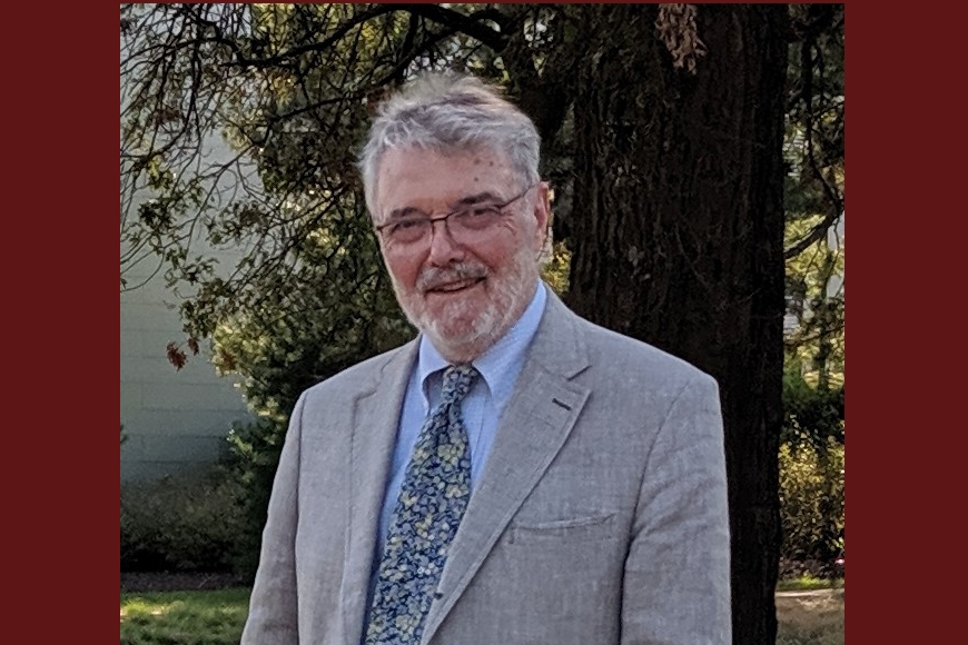person with short gray hair and beard, glasses, light skin, wearing light suitcoat over blue shirt and tie; tree behind