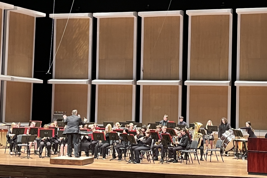 A concert band ensemble actively performing on Ted Mann Concert Hall stage with a conductor standing in front of the ensemble with his hands raised. 