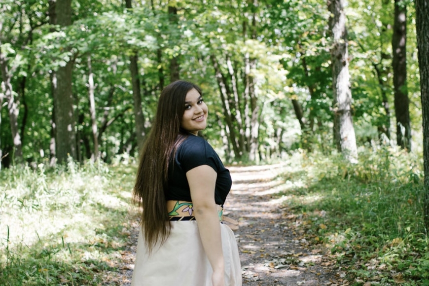 Jaeden King looking back while wearing a blue and white dress, in a sunny green forest